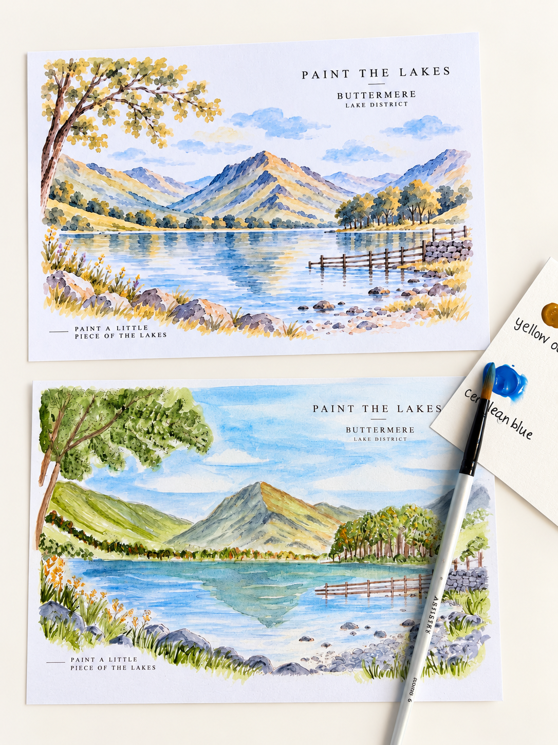 Two printed art cards showing watercolor lake and mountain landscapes, arranged on a white surface with a paintbrush and a small paint palette visible. The cards include the text 'PAINT THE LAKES' and 'BUTTERMERE LAKE DISTRICT.