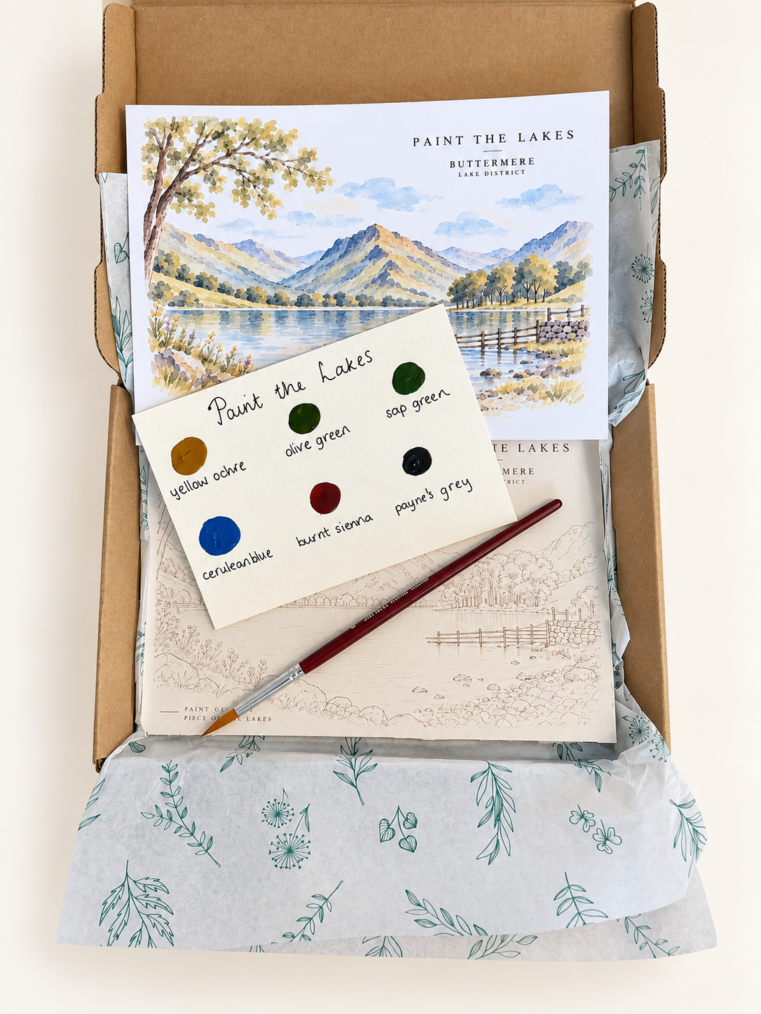 A curated art and craft gift box set from Paint the Lakes, featuring a landscape art print, a paint dot sheet, a fine-tip paintbrush, and a sketchbook nestled in patterned tissue paper inside a cardboard box.