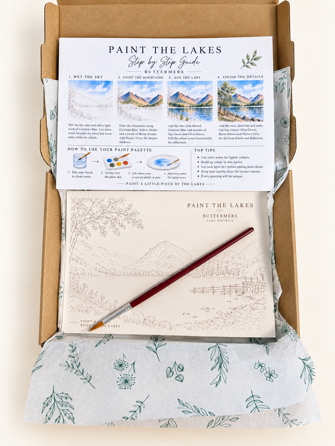 A flat-lay view of a 'Paint the Lakes' step-by-step painting kit inside its cardboard box packaging. The kit includes a printed step-by-step guide, a blank landscape sketch pad, a paintbrush with a reddish-brown handle, and watercolor paint pans nestled in floral-patterned tissue paper.