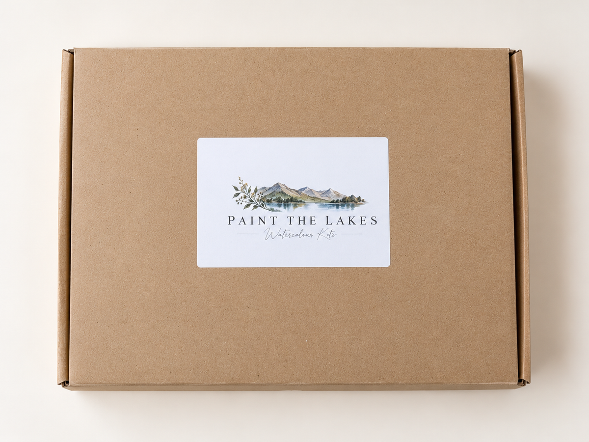 A square brown kraft cardboard box with a white rectangular label on the front featuring a watercolor mountain and lake illustration and the text 'PAINT THE LAKES' and 'Watercolor Kits.